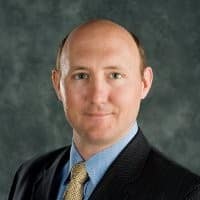 A man in a suit and tie with a blue shirt poses for a professional headshot against a mottled gray background. He has light skin, short brown hair, and a neutral expression.
