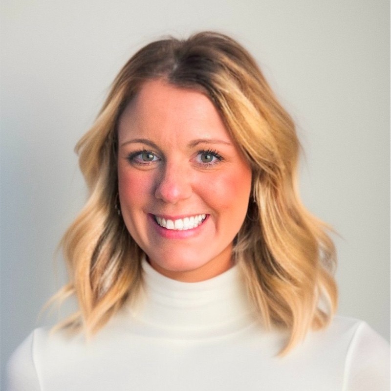 Jennifer Blackburn, a woman with shoulder-length blonde hair styled in loose waves, smiles at the camera. She is wearing a white turtleneck top and is posed in front of a light, neutral background.