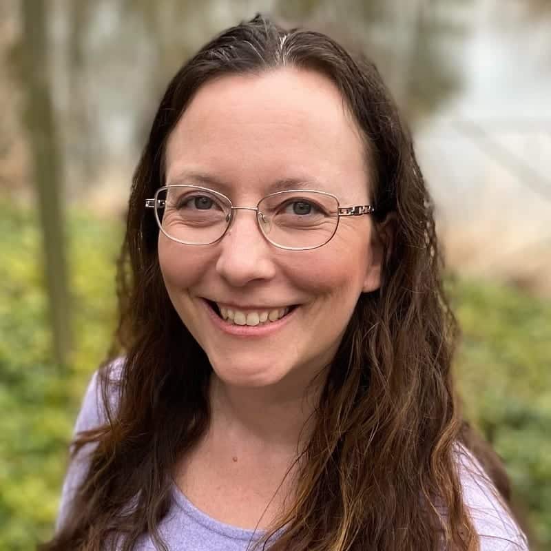 Jillene VanNostrand, a woman with long brown hair and glasses, smiles at the camera outdoors in a light purple top. The background is blurred greenery and trees.