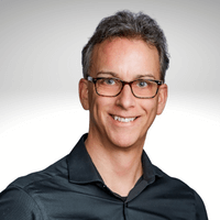 Smiling man with short, gray hair and glasses wearing a dark button-up shirt, posing against a simple light gray background.