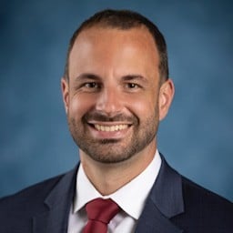 Smiling man with short brown hair and a beard, wearing a dark suit jacket, white shirt, and red tie, posing in front of a blue background—perfect for an Internal Audit professional profile.