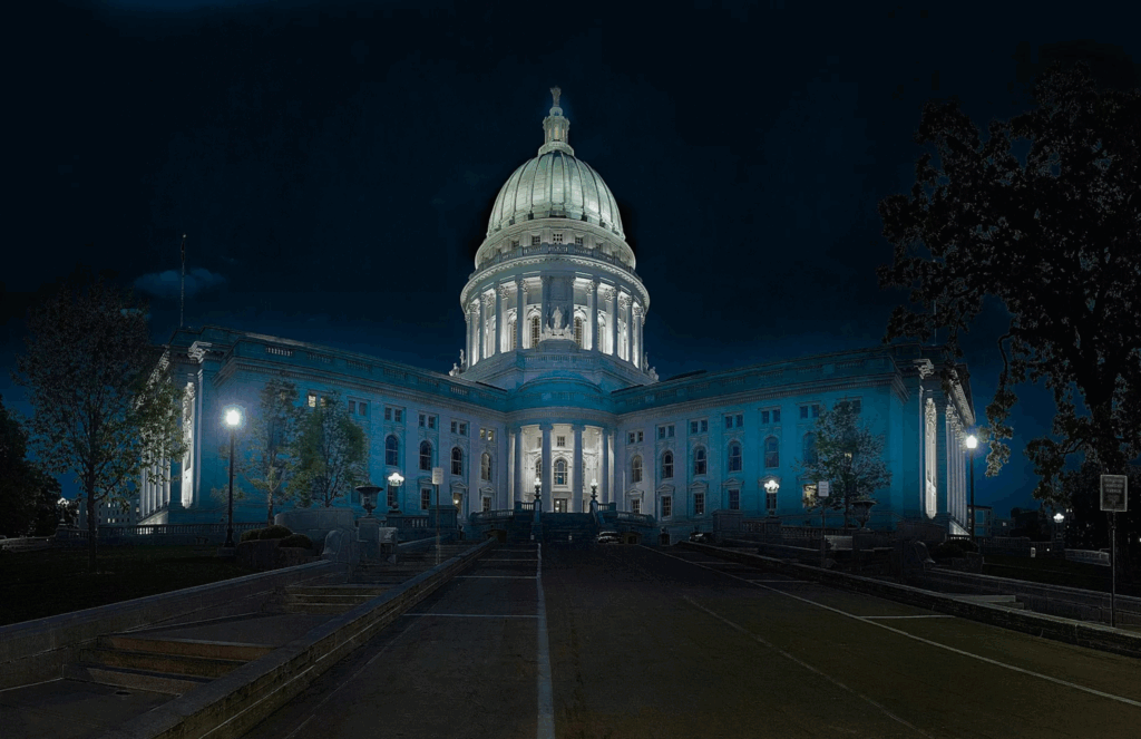 The image shows a large, illuminated domed government building at night, reminiscent of those tied to compliance standards like FedRAMP. Columns and stairs lead up to the entrance, while trees frame the scene beneath a dark sky.