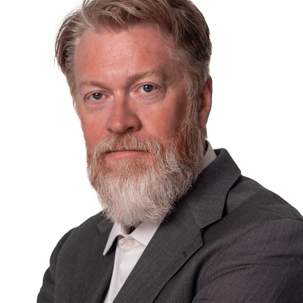 A middle-aged man with light skin, blue eyes, and a full reddish-blond beard wearing a gray suit jacket and white collared shirt, posed against a plain white background.