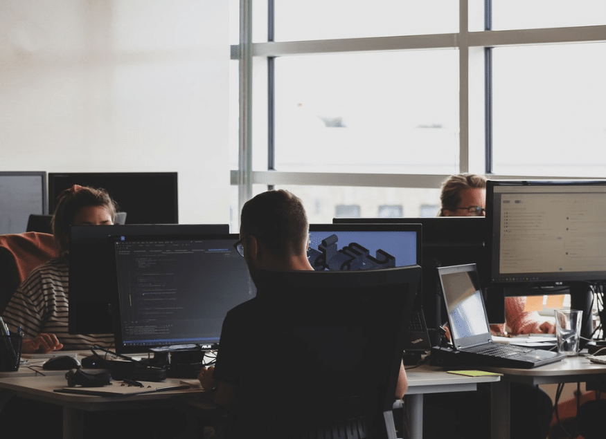Several people are working at desks with computer monitors in a modern, sunlit office space. The collaborative environment suggests a focus on compliance, which is essential for federal contractors adhering to CMMC requirements.