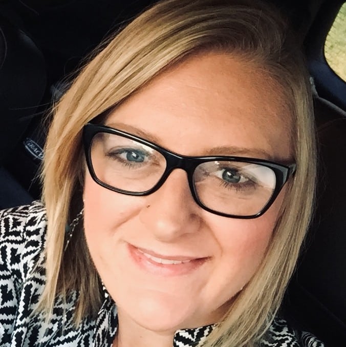Mysti Williams, with blonde hair and glasses, smiles at the camera. She is wearing a patterned top and is seated inside a car, representing Express Employment.