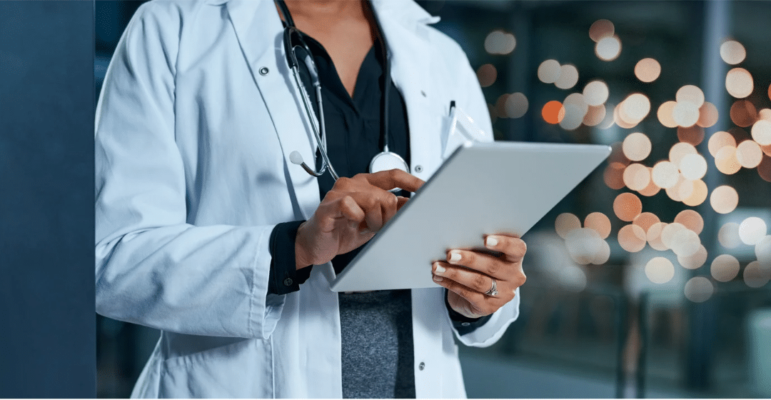 A doctor wearing a white coat and stethoscope uses a tablet device, likely accessing healthcare compliance software, while standing indoors with blurred lights in the background.