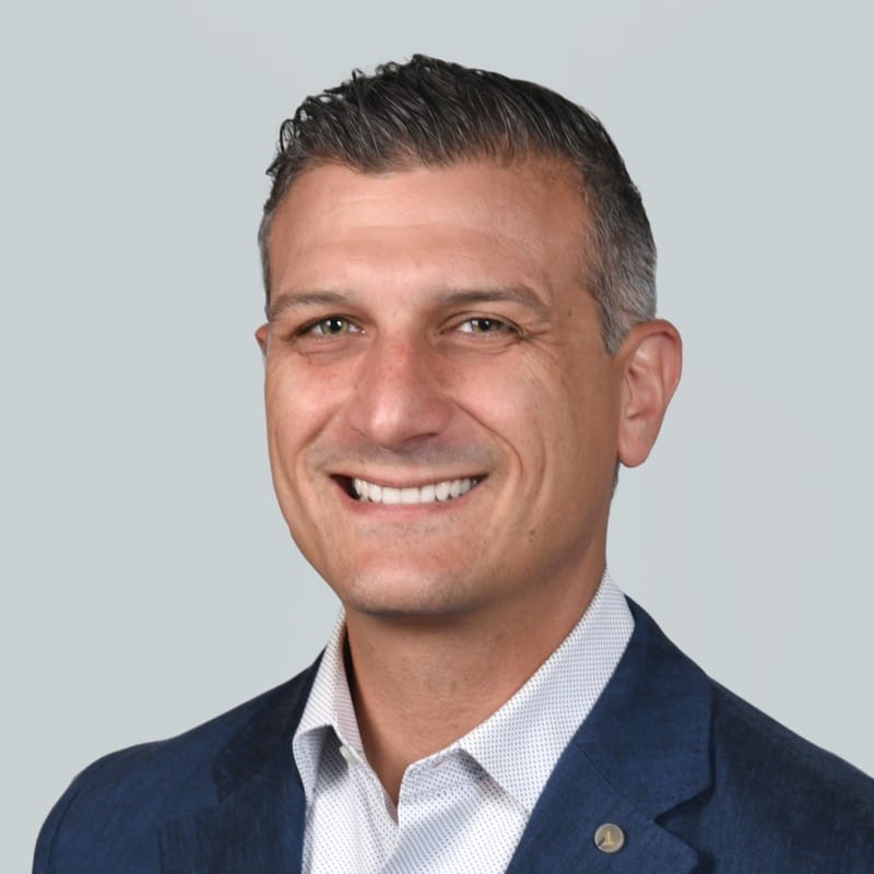 Smiling man with short, dark hair wearing a blue blazer and white dress shirt, posing in front of a plain light gray background—David Johnson of CBRE.