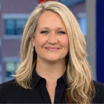 A woman with long blonde hair, wearing a black collared shirt, smiles at the camera. The background is blurred, showing a window with a cityscape outside.