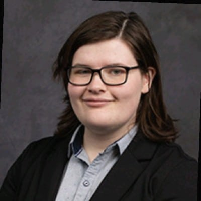 Shay Looney, from Express Employment, stands in front of a dark gray background with straight brown hair, glasses, a light gray shirt, and a black blazer, smiling slightly.