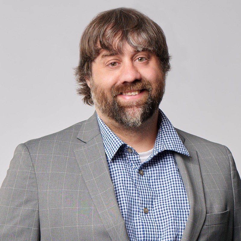 Ryan Lougheed, a man with medium-length brown hair and a beard, wears a grey plaid suit jacket over a blue and white checkered shirt, smiling at the camera against a plain light background.