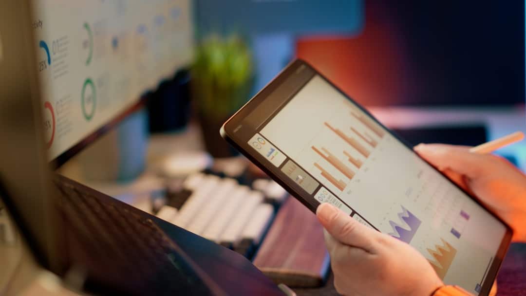 A person holds a tablet displaying bar and line graphs, analyzing performance analytics. A computer monitor with charts and a keyboard are visible on a desk in the background.