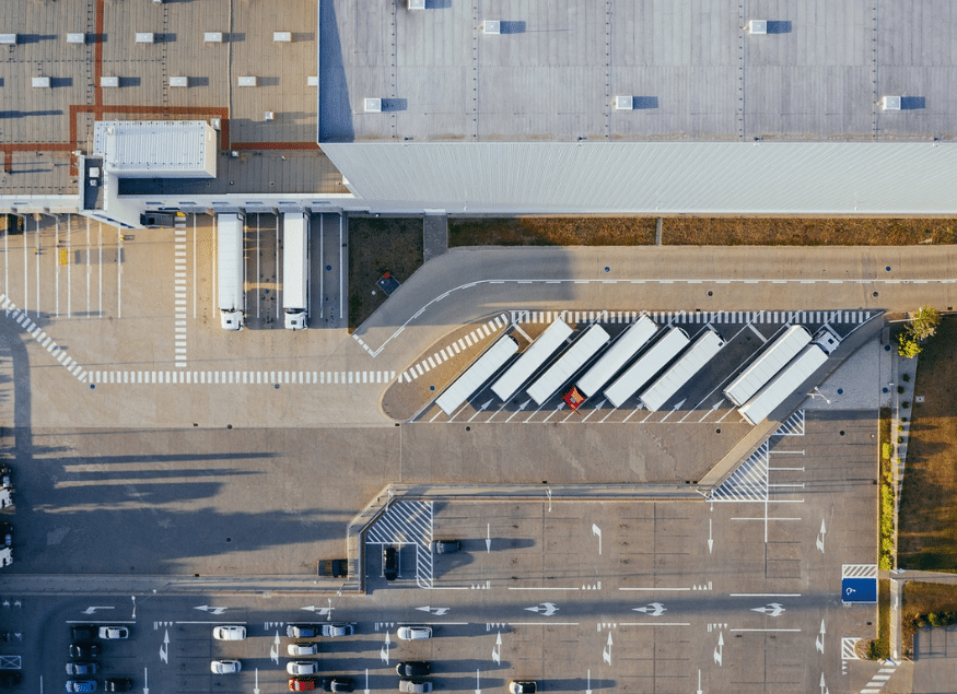 Aerial view of a large warehouse with several trucks at loading docks and cars in a parking lot, showcasing organized lanes and rooftops—an ideal setting for implementing applications of artificial intelligence in logistics and operations.
