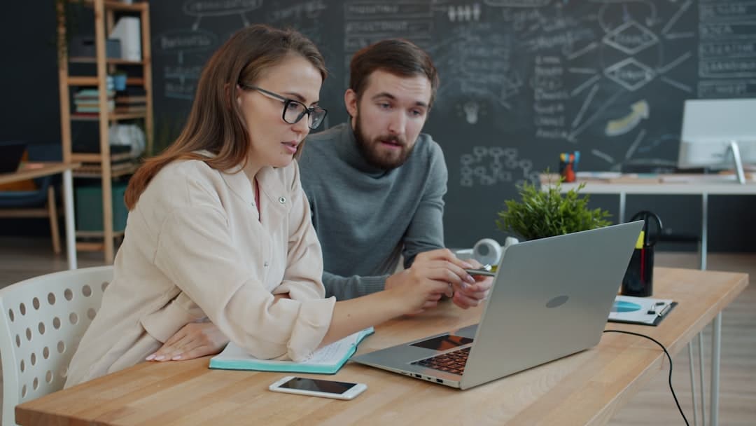 Two people sit at a desk, looking at a laptop screen and discussing alternative business solutions. The desk holds a notebook, pen, smartphone, and small potted plant. A chalkboard with diagrams and writing is visible in the background.