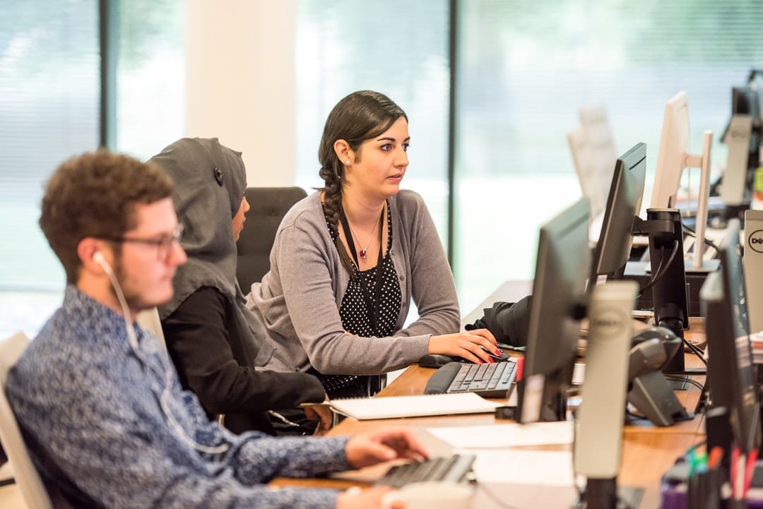 Three people sit at a long desk working on desktop computers in an office setting, focusing on a woman typing. They appear engaged in customer care and services, with one wearing a hijab and another with glasses and earphones.