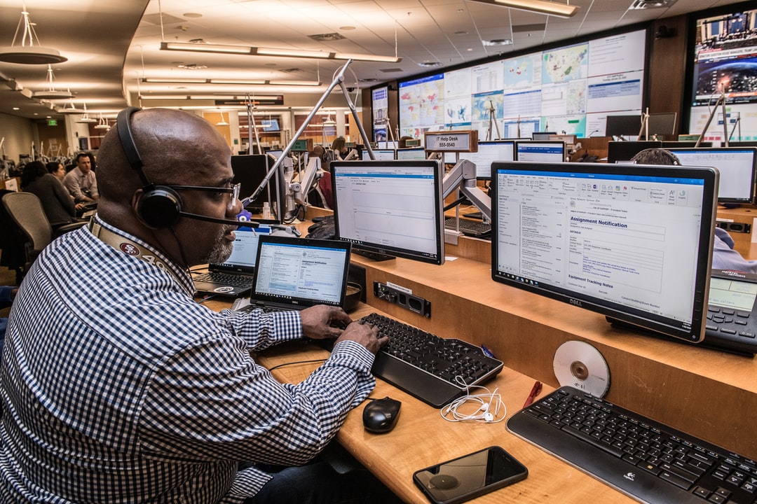 A man wearing headphones works at a desk with multiple computer monitors and a laptop in a busy control room, providing help desk solutions amid people, large screens, and workstations.