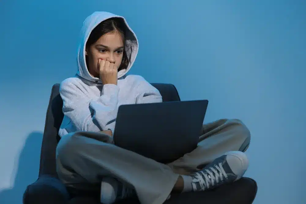A young woman in a hoodie sits on a chair, focused on her laptop, with a neutral background.